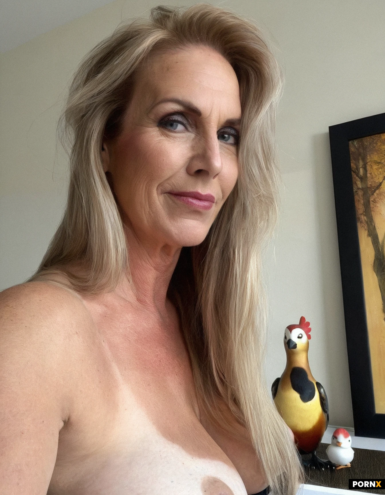 portrait of an adult woman in her early fifties, long straight blonde hair brushed slightly off-center, fair skin marked by fine lines, mild sagging at the jawline, neck and under the eyes, and a soft closed-mouth smile; her green eyes gaze gently forward. She is completely nude, her breasts visibly deflated and sagging, resting naturally against her torso, while the scene is set against a plain white wall with a large framed landscape painting of trees behind her and a white dresser to the side holding a colorful bird figurine and a framed family photo. Soft, diffused natural indoor lighting creates subtle rim lighting along the hair and a shallow depth of field that blurs the background, emphasizing realistic skin texture, age-related imperfections and the delicate interplay of light on sagging flesh. Captured from a slightly lower eye-level angle to accentuate her posture, the composition evokes a high-detail photographic style reminiscent of fine-art portraiture. Full body