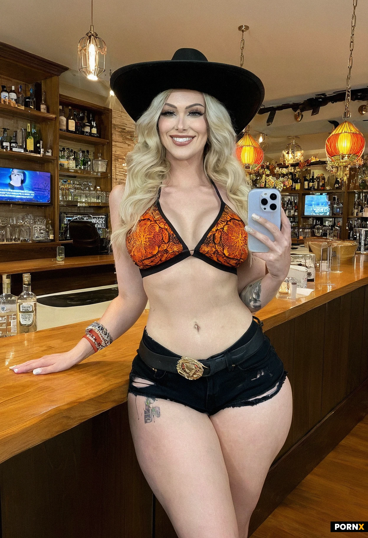 A full-length vertical social-media screenshot shows a smiling adult woman with light skin and a curvy, toned build, standing near the center of a brightly lit bar or salon interior. She faces the camera with a relaxed, slightly angled stance, one hand near her hip and the other lowered beside her thigh. She has long, wavy blonde hair, softly made-up features, and wears a black cowboy hat with an orange patterned brim and decoration. Her outfit includes a colorful orange, red, and black patterned bikini-style top, black low-rise shorts with a belt and dangling metal details, a black wristband, and a red band around her other wrist. Shelves, bottles, counters, and colorful illuminated displays fill the busy background. The framing is a slightly low, close vertical phone-camera view, with warm overhead lighting and vivid accents. Social-media interface elements, including a search bar and reaction icons, overlay the image. The scene has a casual, nightlife atmosphere and a candid influencer-video aesthetic.