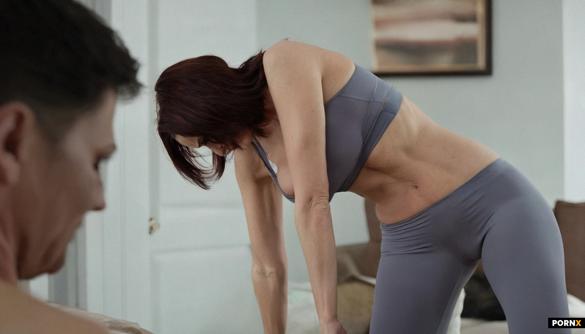 A warm-lit living room frames a flabby mother in her early thirties. tight blouse, tight pants. She has small freely sagging breasts in a dangling décolletage. Her posture is hunched forward, shoulders turned inward, creating a taboo, teasing tableau of vulnerability. The viewing angle sits at a slightly low angle from behind, capturing a close-up with shallow depth of field that isolates her flabby butt, while the muted décor blur into a cozy ambiance. Rendered in ultra-realistic DSLR quality with crisp detail for a cinematic, intimate maternal candid feel.