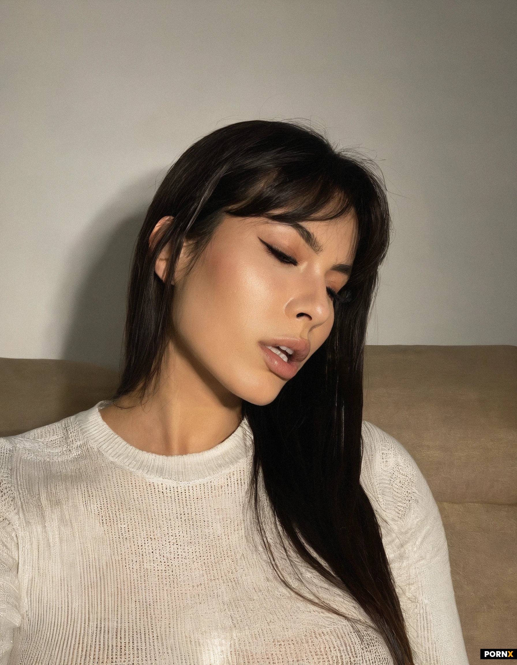 Close-up side-profile portrait of a latina woman, mid-20s, seated indoors. She faces left, head tilted slightly upward, mouth open with lips parted and tongue visible. A thin, translucent white strand stretches from the corner of her lower lip toward the left edge of the frame, catching the light. Dark brown, slightly damp-looking hair with wispy bangs falls across her forehead and cheek; longer hair hangs past her shoulders. Fair skin with a soft sheen, defined eyebrows, long lashes, a small straight nose, and glossy lips. She wears a black knit sweater with a white crew-neck undershirt visible at the collar.
Warm indoor lighting from the front-left highlights her cheekbone, nose bridge, and lower lip, with gentle shadows under the jaw and along the neck. Background is a softly blurred beige sofa and neutral wall. Shallow depth of field, phone-camera look, slight motion softness in the hair strands.