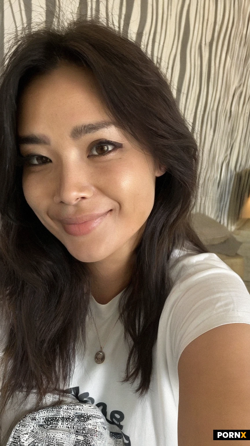 A realistic high-angle selfie of a smiling 27woman with long, slightly wavy dark brown hair and light freckles on her face. She is sitting on a bed with white sheets and a wooden headboard behind her. She wears a simple white short-sleeve t-shirt with a black graphic print on the front and ultra-short light blue denim shorts. A delicate silver pendant necklace is around her neck. The perspective is a first-person point-of-view selfie, with her right arm extended toward the camera, creating a natural foreground blur. The background features light-colored vertically striped wallpaper. Casual indoor amateur photography style, direct flash lighting, sharp focus on her face, natural look. --ar 2:3 --v 6.0