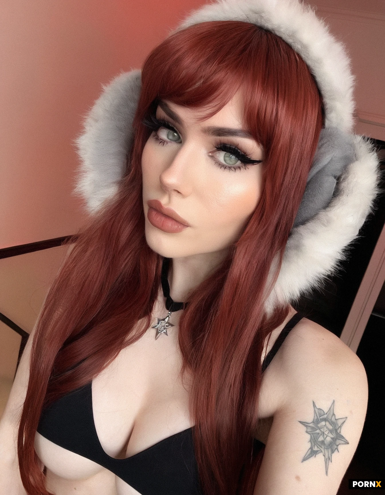 Photorealistic selfie portrait of a sexy adult woman with pale fair skin, long copper-red hair with straight wispy bangs, light green-gray eyes, subtle winged black eyeliner, long eyelashes, softly arched eyebrows, and natural pink lips. She has a slim build and visible black-and-gray tattoos, including a prominent heart-shaped tattoo centered on her upper chest and small tattoos around her shoulders and collarbone. She is wearing a black sleeveless low-cut top and a black furry winter trapper hat with ear flaps, decorated with a small red-and-white star-like pin on the front. She is looking directly into the camera with a calm, slightly sultry expression. Close-up casual smartphone selfie, head and upper torso, slightly above eye level, natural indoor lighting, warm neutral room in the background, shallow depth of field, realistic skin texture, individual hair strands, authentic smartphone photography, candid Instagram-style composition, high detail, photorealistic, 4:5 portrait aspect ratio.